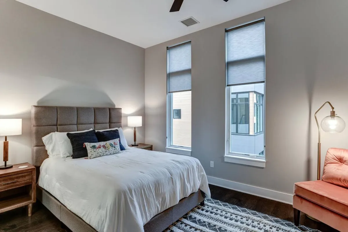 A serene bedroom featuring a large, inviting bed flanked by nightstands and windows that let in abundant natural light. An accent chair by the window and soft textures create a peaceful and restful environment.
