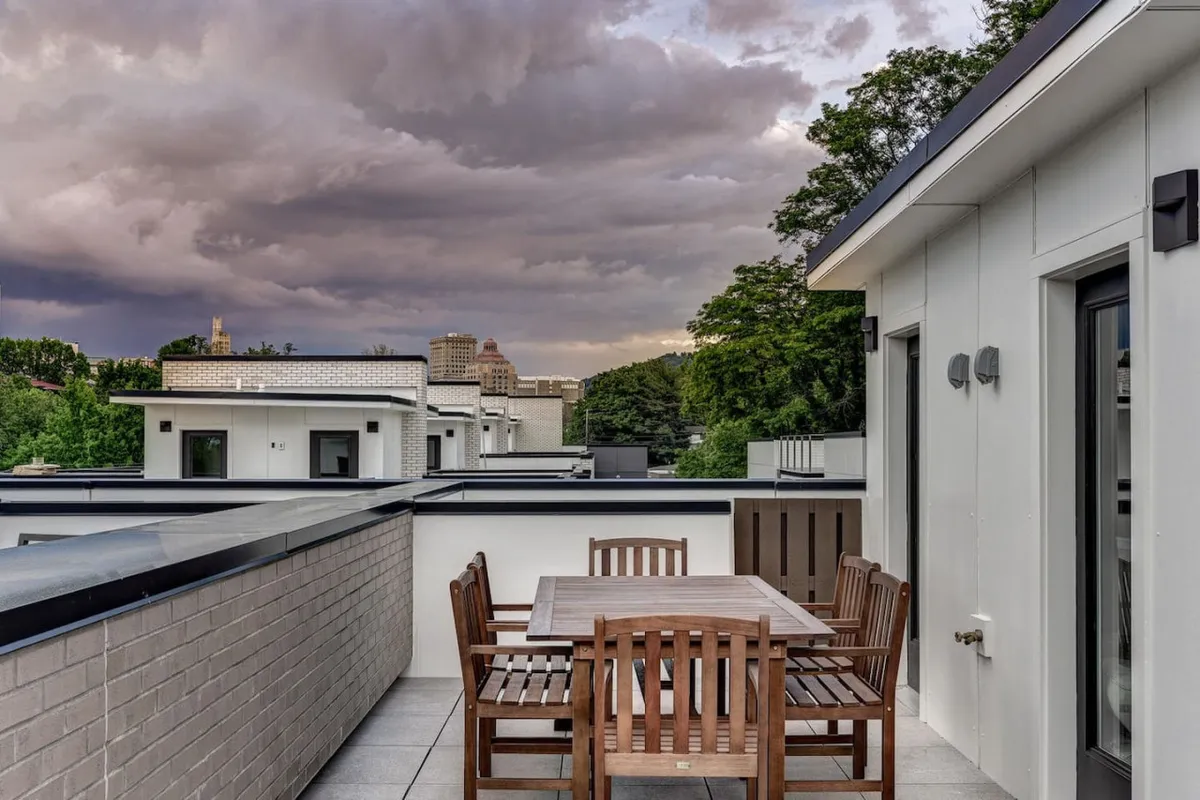 This exterior shot showcases a wooden dining table set amid surrounding greenery and under a cloudy sky. The setting suggests opportunities for outdoor dining, morning coffee, and leisurely gatherings with the city as a distant backdrop.