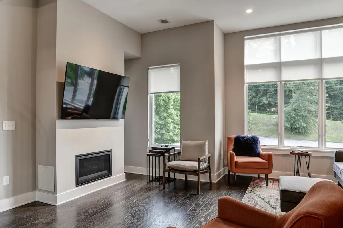 This image showcases a cozy, modern living area with a large television above a warm fireplace, surrounded by plush seating. Expansive windows let in natural light and offer a glimpse of the outdoors, creating an inviting space perfect for relaxing, socializing, or enjoying a quiet evening.