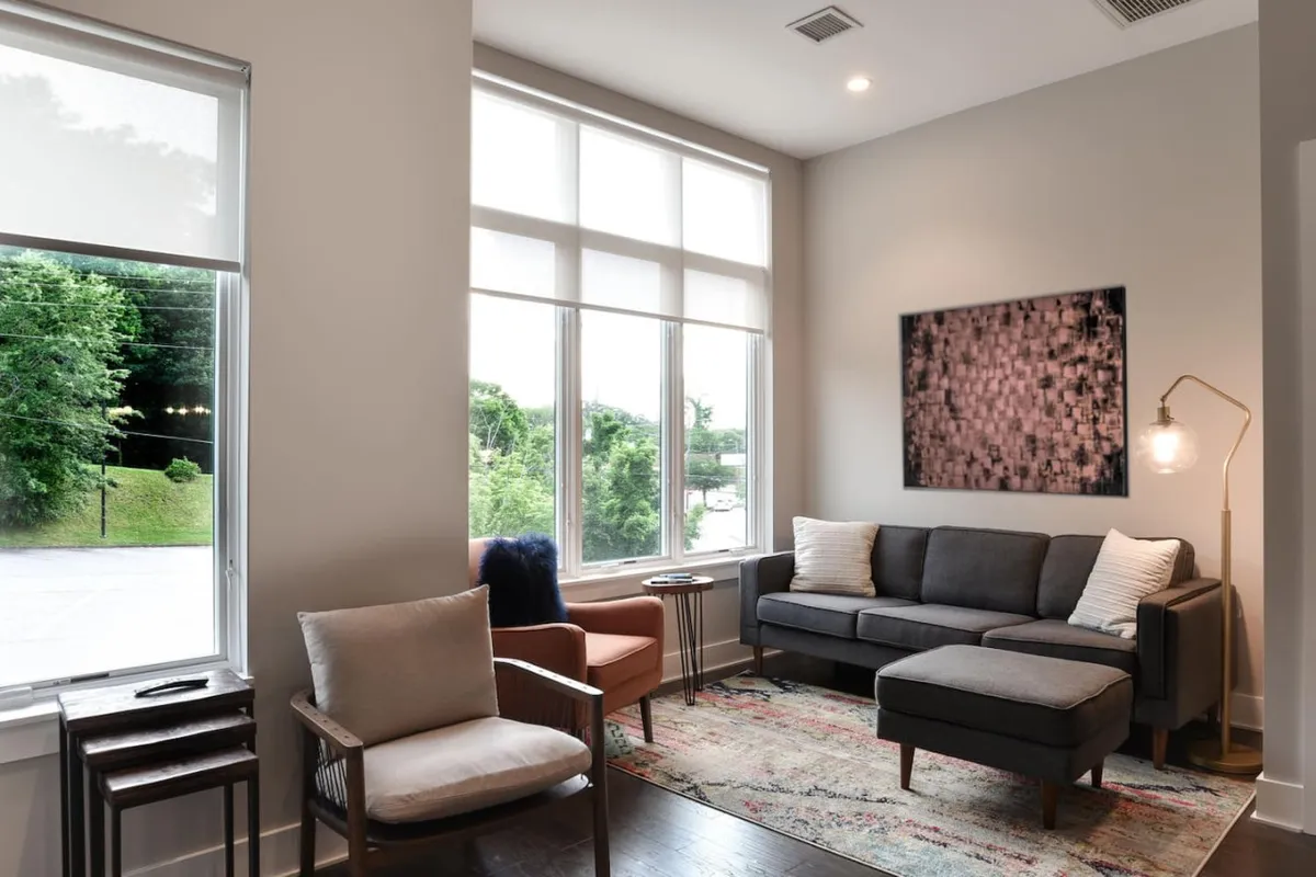 A cozy living room filled with natural light from large windows, featuring a comfortable sofa, vibrant artwork, a decorative rug, and warm ambient lighting. The arrangement encourages relaxation, conversation, and togetherness.