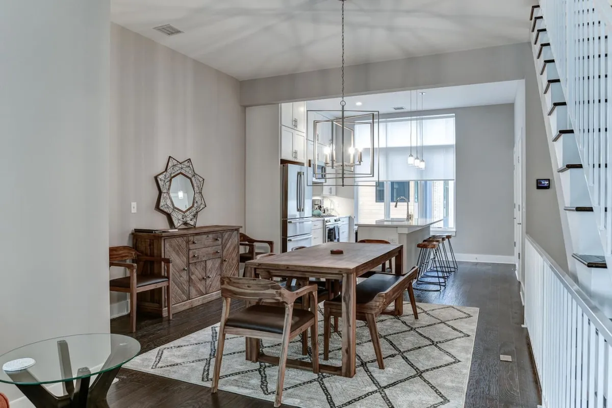 A spacious, inviting dining area with a large table set for a meal, illuminated by a striking chandelier. Reflections in the decorative mirror add depth, while an open view to the kitchen hints at easy movement between spaces. The setting feels accessible and welcoming, suitable for family gatherings or entertaining guests.