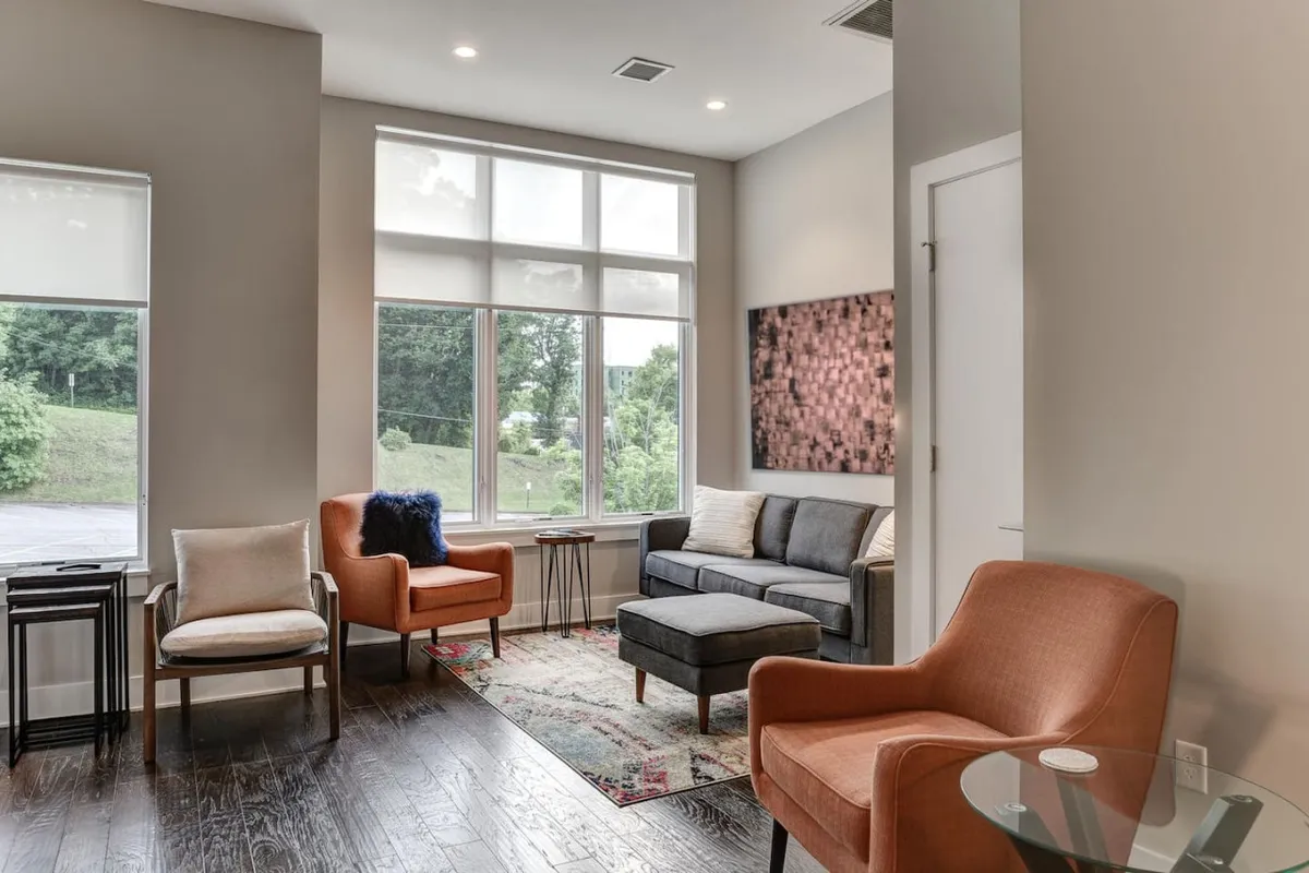A bright and modern living space with large windows letting in natural light, colorful artwork on the wall, and a stylish seating arrangement set up for socializing or relaxing. The layout invites guests to gather comfortably and enjoy the ambiance.