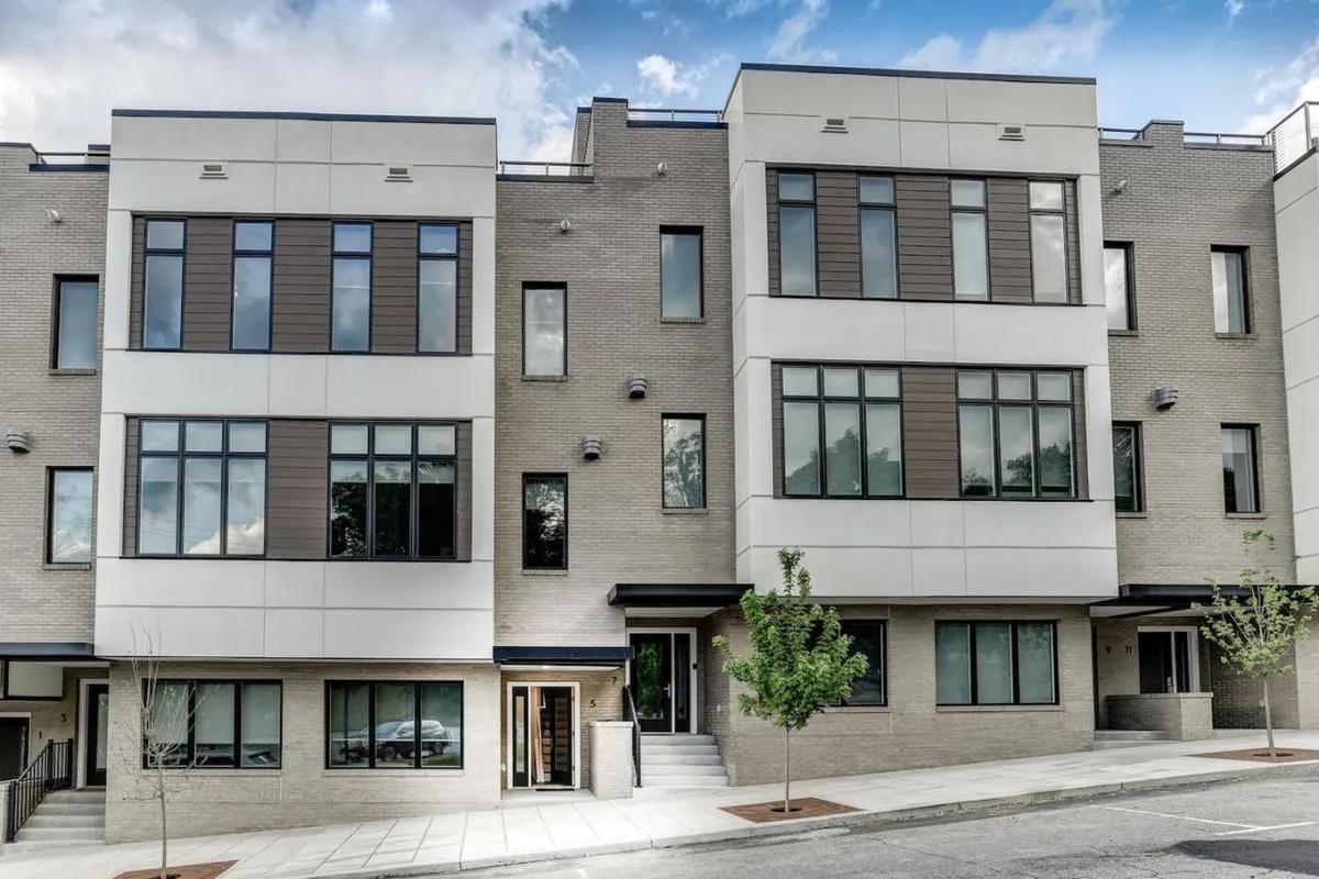 The image captures the exterior of the townhome, showcasing its modern architecture with large windows, a symmetrical design, and a landscaped entrance. The setting is welcoming and contemporary, with hints of community activity and the possibility of outdoor relaxation. The visual focus on the entrance hints at easy access to the vibrant South Slope and downtown Asheville scene just beyond.