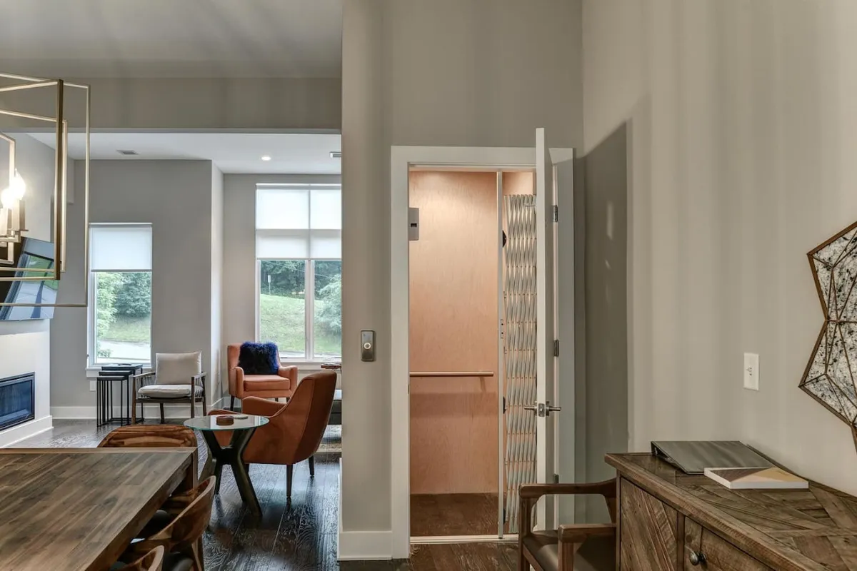 The image displays an open-plan dining and living area filled with natural light. The elevator door is visible in the background, indicating easy access to upper floors. Comfortable seating and large windows make the space feel modern and accessible.