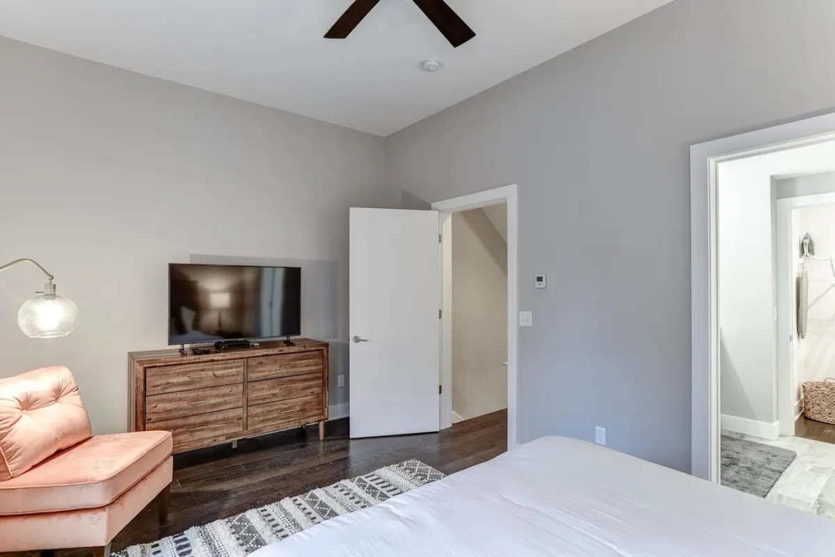 A cozy, sunlit sitting area with a bright orange armchair, a sleek wooden TV stand, and a glimpse of a neatly made bed. The open door and natural light streaming in create a welcoming transition between spaces, suggesting freedom and ease of movement.