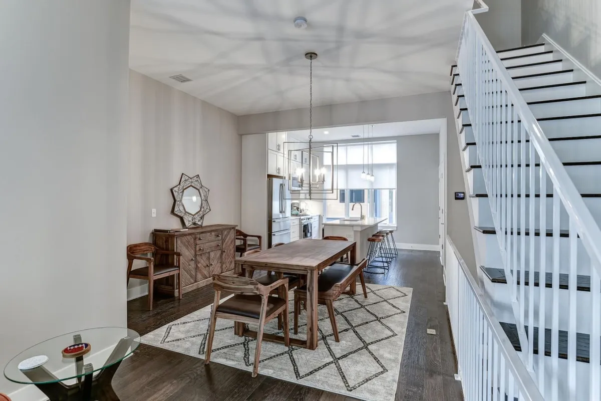 A spacious dining area featuring an elegant dining table, a sparkling chandelier above, a decorative mirror, and a staircase in the background. The setting is both inviting and sophisticated, suitable for hosting group dinners or celebrations.